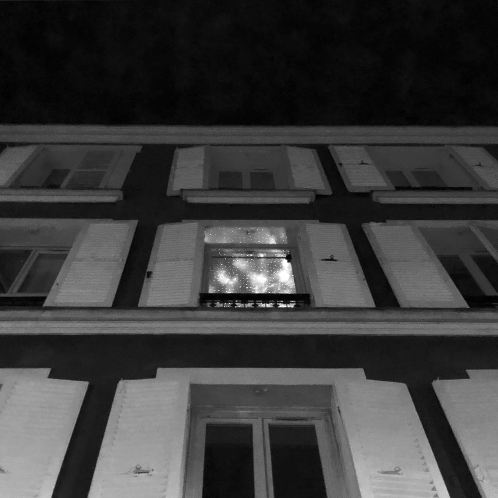 De nuit, immeuble avec tous les volets fermés, sauf une fenêtre d'où émanent des lumières de fête