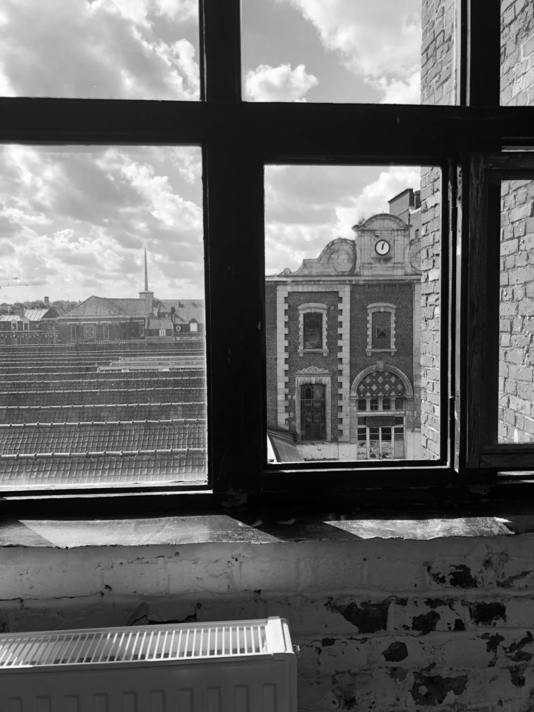 Photo en noir et blanc à travers une fenêtre : vue sur le fronton d'une ancienne usine stylée mais délabrée et les toits