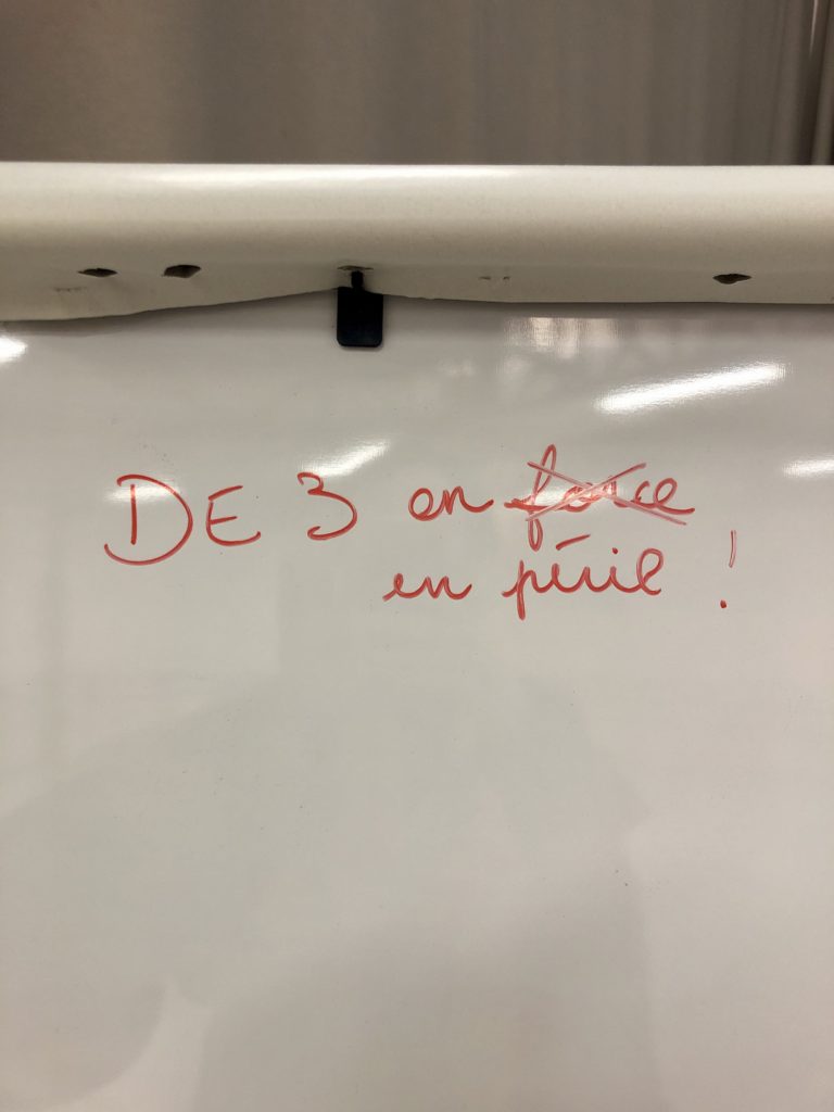 Tableau blanc sur lequel il est écrit au feutre rouge "DE3 en force", avec "force" barré et remplacé par "en péril"