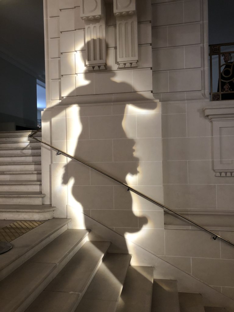 Ombre d'une statue cernée d'une aura lumineuse, projetée sur le mur derrière un escalier