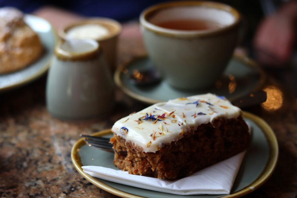 Carrot cake avec sa couche de sucre glace décorée de fleurs, et en arrière-plan flou, tasse de thé et assiette avec scone