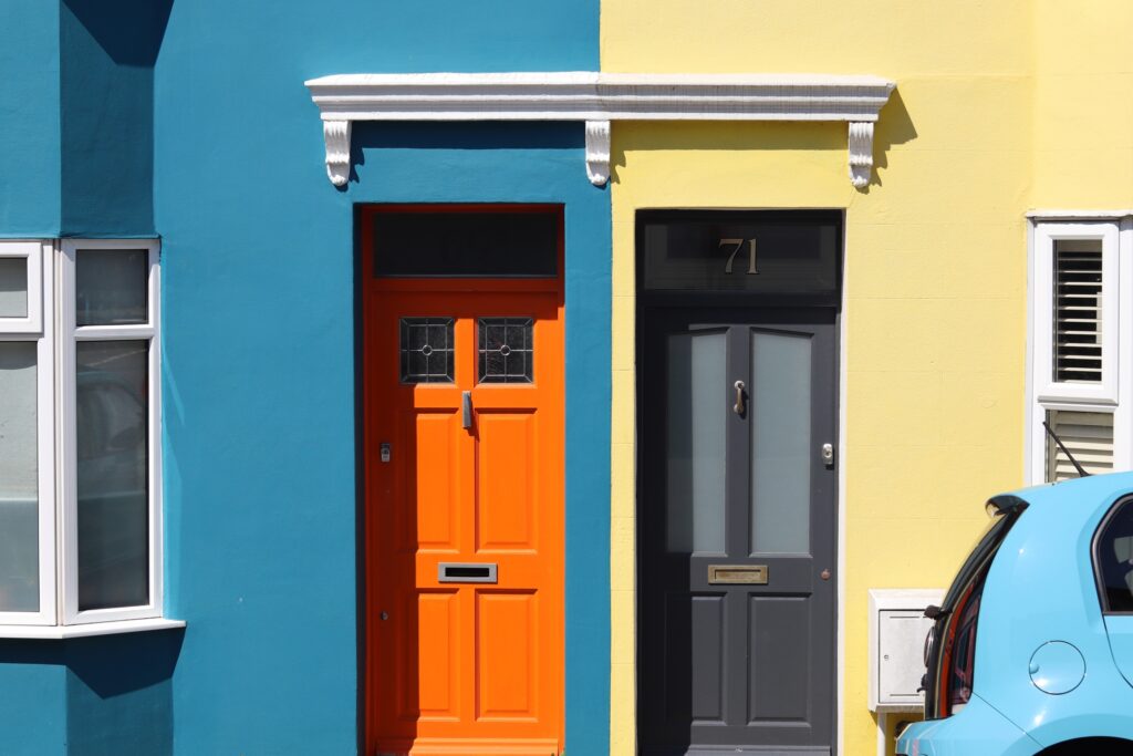 Séparation colorée de deux maisons mitoyennes : une bleu avec porte orange et une jaune avec porte gris sombre. Devant la seconde est garée une voiture bleu pastel qui fait écho à l'autre maison