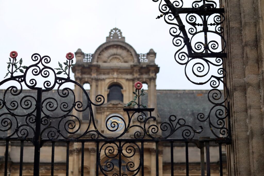 Bâtiment à fronton et colonnes flou derrière un premier plan de grilles avec des volutes et des fleurs en fer forgé.