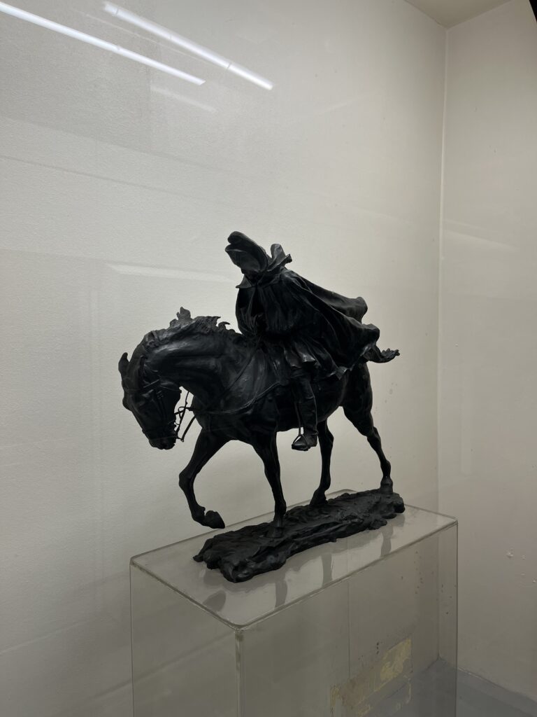 Une sculpture d'un homme à cheval en cape, qui lutte contre le vent (impression renforcée par le reflet des néons dans la vitre)