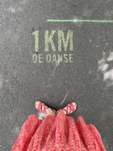 Jupe rouge et sandales assorties. Pieds en première position pris en plongée à côté d'une inscription en jaune-vert au sol "1 km de danse"