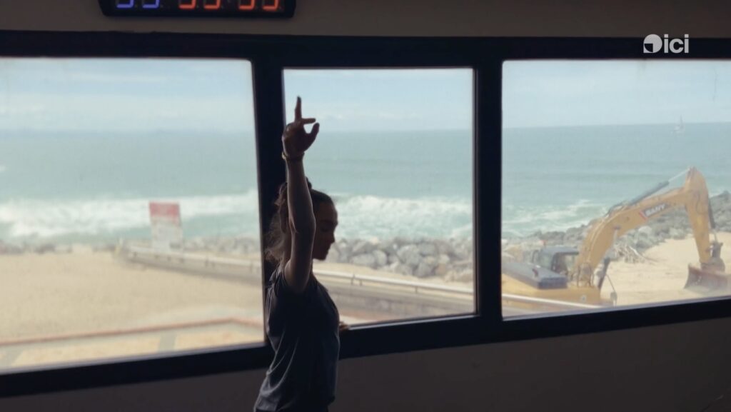 Mathilde à la barre devant des fenêtres où l'on aperçoit une plage, la mer et un tractopelle